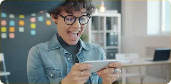 Une jeune femme portant des lunettes et une chemise en jean sourit avec enthousiasme et ouvre grand la bouche de joie en regardant son smartphone qu'elle tient à deux mains dans un bureau lumineux.