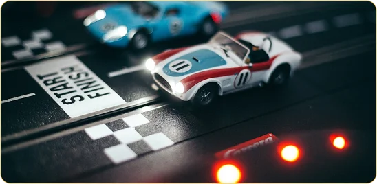 Deux voitures de course miniatures aux phares allumés franchissent la ligne d'arrivée sur un circuit de jeu sous une lumière tamisée, recréant l'atmosphère intense d'une véritable compétition automobile.